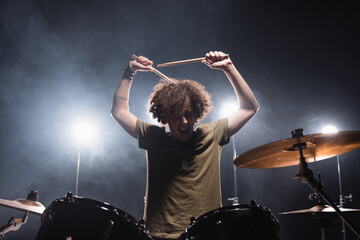 Curly musician shouting, while holding drumsticks and sitting at drum kit with backlit on background