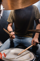 Obscure face of musician with drumsticks playing drum on black