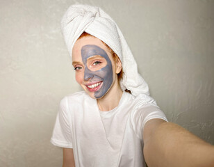 portrait of a happy young woman with a clay mask on her face, and a towel on her head is photographed on a smartphone, selfie.