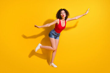 Fototapeta na wymiar Full size photo of lovely dreamy young girl raise hands leg lips send air kiss attract guys party wear red singlet unclothed shoulders denim shorts isolated vibrant yellow color background