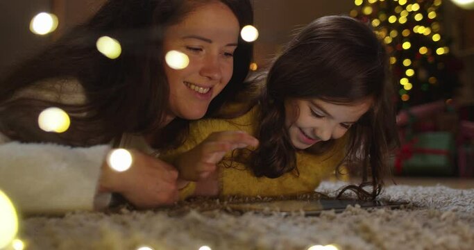 Close up of joyful cute Caucasian little girl with mom laying on floor in decorated home browsing and tapping on tablet together on Christmas Eve. Family time. Kid with mother using device on New Year