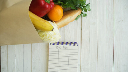 Full paper bag of food products with blank little notebook on wooden table, top view