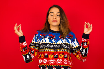 Young beautiful Caucasian woman wearing Christmas sweater against red wall, doing yoga, keeping...