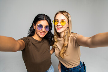 Portrait of two lovely women in sunglasses standing and taking a selfie isolated over white background