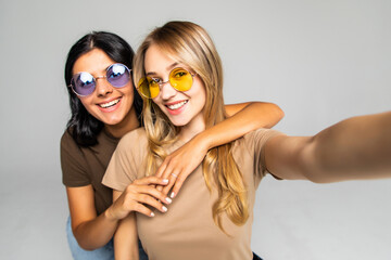 Portrait of two lovely women in sunglasses standing and taking a selfie isolated over white background