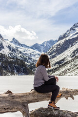 Naklejka premium Beautiful young woman sitting next to stunning winter landscape. Concept of travel and adventure at the frozen Gaube Lake (Lac de Gaube) in the French Pyrenees, Cauterets, France.