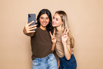 Portrait of two smiling women using smartphone while gesturing peace sign isolated over beige background