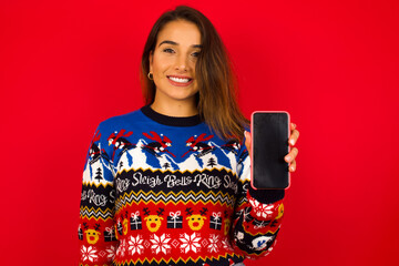 Smiling young beautiful Caucasian woman wearing Christmas sweater against red wall,  showing  empty phone screen. Advertisement and communication concept.