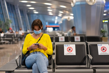 A beautiful woman is waiting for her flight, sitting in the airport lounge in a protective mask with a phone in her hands.