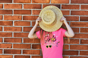 Girl covering her face with a straw hat on a brick wall background.