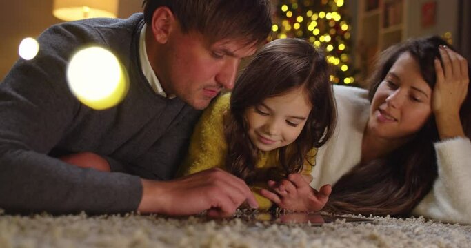 Close up of Caucasian joyful mom and dad with little cute daughter on floor in decorated home typing and browsing on tablet choosing xmas gifts together on New Year Eve. Family time