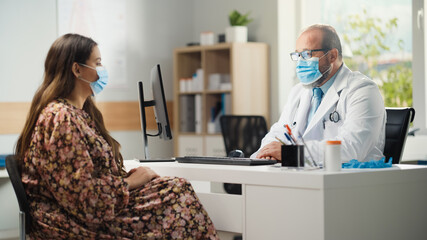 Hispanic Doctor's Office: Physician Sitting at His Desk Talks With Sick Female Patient. Medical Health Care Specialist Giving Advice, Prescribing Medicine, Recommending Treatment. Both Wear Face Masks