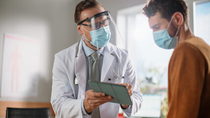 Doctor Consultation Office: Professional Physician Talks to the Male Patient, Uses Digital Tablet Computer to Explain Test Results, Treatment Plan, Prescribes Medicine. Wearing Face Masks and Visor.