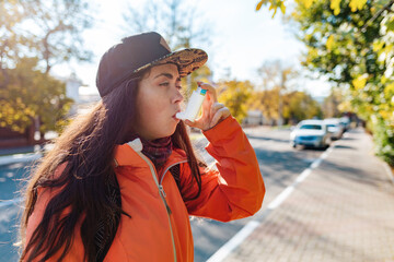 Chronic disease. A teenage girl in casual clothes and cap using an inhaler on the street, having an...