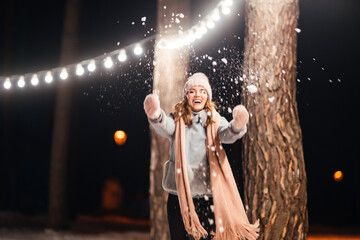 Beautiful young woman Playing with snow evening in the winter park. Happy  woman having fun on winter holidays. Christmas, New year. Winter lifestyle.