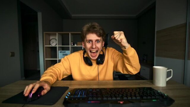 Joyful gamer sits at home in the bedroom at the computer and rejoices in victory with a happy face. young man wins a computer game, looks at the screen and shows a fist with a happy face.