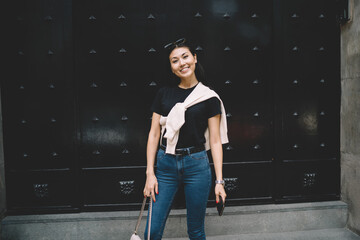 Half length portrait of trendy dressed hipster girl standing on street in city enjoying free time looking at camera, smiling asian young woman in stylish wear posing on urban setting background