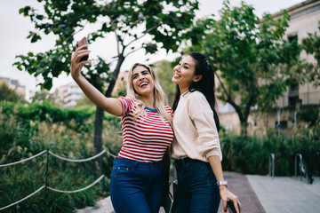 Happy multiracial female colleagues positing for selfie resting together on urban settings enjoying meeting, cheerful hipster girl making photo on smartphone camera share content in social networks