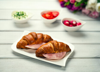 Fresh croissant sandwich with ham, tomatoes and radish on white table. Healthy romantic breakfast for two with bouquet in the background