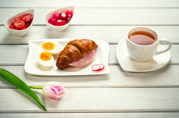 Fresh croissant sandwich with ham, eggs, tomatoes and radish and mug of tea on white table. Healthy romantic breakfast served with love and pink tulip flower in foreground