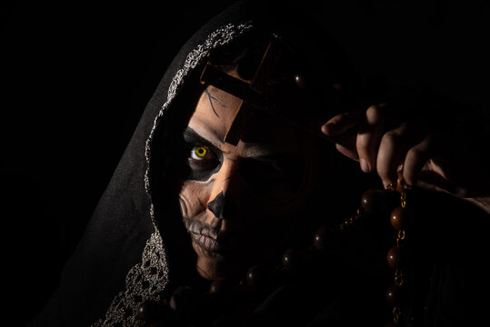 The death. Artistic makeup representing death, Low Key image, Death holding a wooden crucifix, selective focus.