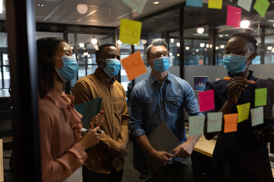 Diverse office colleagues wearing face masks discussing over memo notes on glass board at modern off