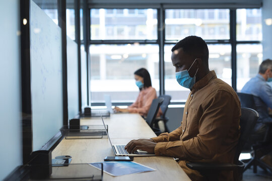 African american man wearing face mask using laptop while sitting on his desk at modern office