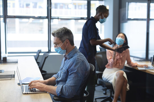 Caucasian man wearing face mask using laptop while african american woman and asian woman wearing fa