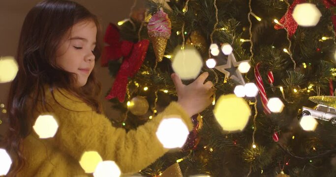Close up portrait of happy cute little Caucasian girl at home decorating christmas tree alone, looking at camera and smiling on New Year. Christmas preparation. Festive period. Twinkle lights