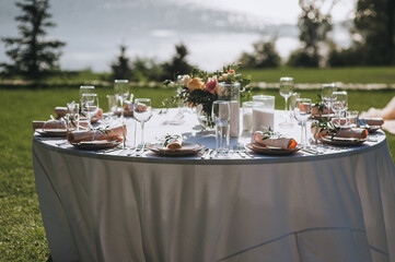 A large round table with a white tablecloth and dishes, a bouquet of flowers stands on the green grass in the park in nature. Wedding ceremony, party. Holiday decorations.