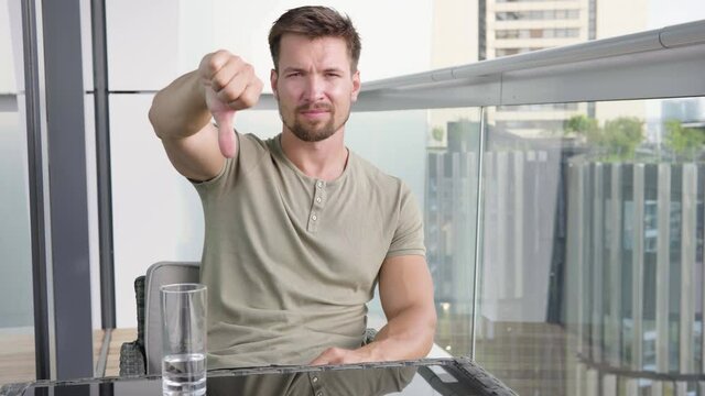A young handsome man shows a thumb down to the camera and shakes his head as he sits on a balcony in a luxurious apartment