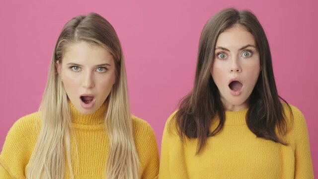 Close-up view of a two shocked women are looking to the camera standing isolated over pink background in studio