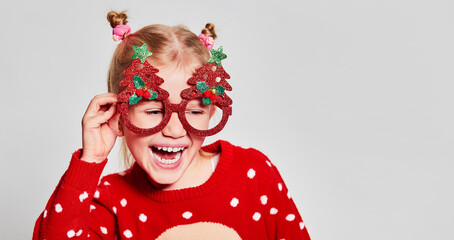 Funny blond little girl wearing red christmas sweater and party glasses, making funny faces on gray background with copy space. Christmas mood concept.