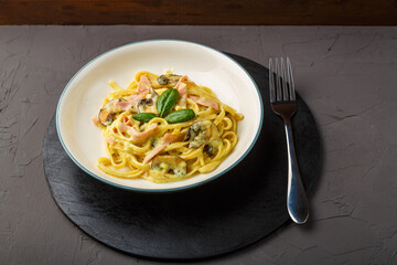 a plate of pasta with ham and mushrooms in a creamy sauce with basil on a black round stand with a fork on a gray background.