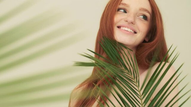 A beautiful woman is posing to the camera while holding a palm leaves near her face standing isolated over beige background in studio