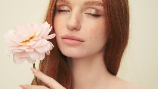 A close-up view of a calm nice woman is posing to the camera while holding a flower standing isolated over beige background in studio