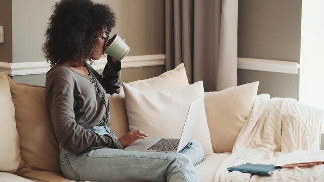 Young positive happy african woman sitting indoors at home and using laptop computer while drinking coffee