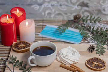 Fototapeta na wymiar Medical mask, Christmas candles, coffee mug on a wooden table, air meringue, dried orange, cinnamon and accessories for winter holidays. New Year