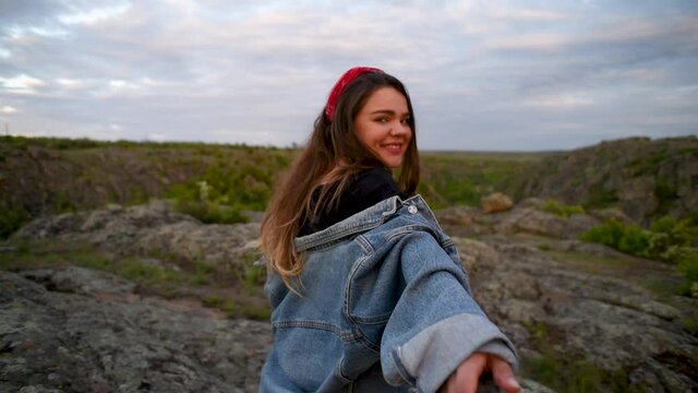 Smiling Brunette girl with a hairband and jeans jacket invites to follow her in the background of canyon