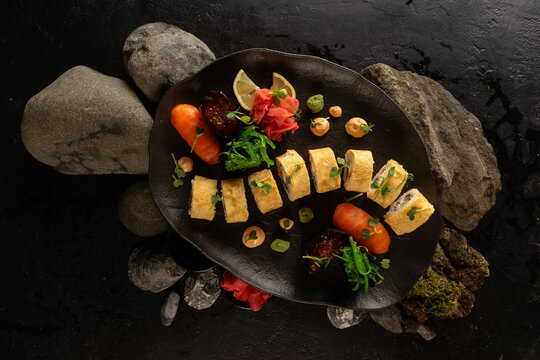 A set of sushi on a beautiful black plate. Japanese dish. Sushi rolls with Japanese tamago amlet and salmon sashimi with rice, gunkans with red caviar, ginger, wasabi, chuka salad and peanut sauce.