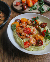 Glazed shrimp with garlic and honey sauce over creamy mashed potatoes and topped with colourful cherry tomatoes and coriander leaves. Beautiful spring meal plated on wooden table. 