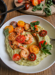 Glazed shrimp with garlic and honey sauce over creamy mashed potatoes and topped with colourful cherry tomatoes and coriander leaves. Beautiful spring meal plated on wooden table. 