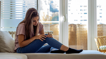 Photo of beautiful young woman sitting on a sofa in the living room at home and touching her knee...