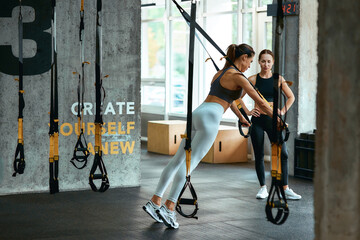 Young athletic woman in sportswear exercising with trx fitness straps with personal trainer at gym, doing push ups