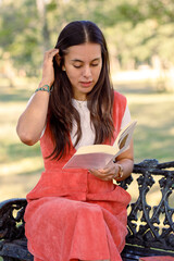 Mujer latina joven, leyendo un libro sentada en una banca de un parque, estudiando al aire libre.