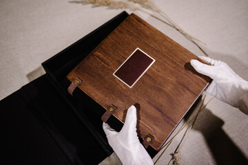 The hands of a man in white gloves hold a photo book with a wooden designer cover and leather...