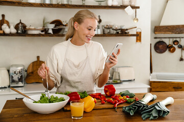 Beautiful happy woman using cellphone and smiling while making lunch