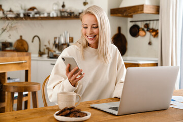 Beautiful happy woman using smartphone while working with laptop