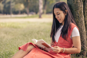 Chica leyendo un libro en el parque, sentada en el césped recargada de un árbol. 