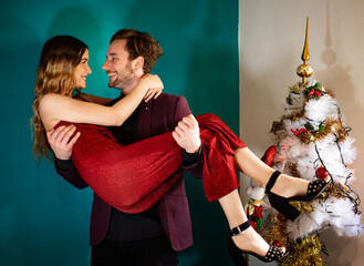 Boyfriend holding his girlfriend in the arms while they wear fashionable outfit during Christmas.
Young couple in fashionable outfit with christmas tree in the background.